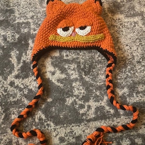 May include: An orange crocheted hat resembling Garfield the cat. The hat features white eyes, a yellow mustache, and black accents. Braided orange and black cords with tassels hang from the hat.