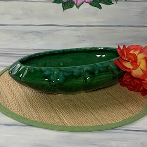 May include: A glossy, emerald green ceramic planter with a ribbed design, resting on a round, woven mat with a green border. A cluster of orange and yellow roses sits to the right. The background features a floral painting.
