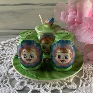 May include: A set of four green and blue ceramic salt and pepper shakers shaped like flowers with smiling faces. The shakers are on a green and white ceramic plate.