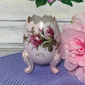 May include: A light pink, egg-shaped ceramic vase with a scalloped rim and three small feet. The vase is decorated with a floral pattern of red roses and green leaves. Gold trim accents the rim and feet. The vase is a decorative item.