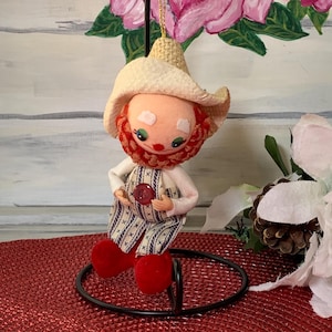 May include: A whimsical Christmas ornament featuring a doll-like figure with a straw hat, red beard, and overalls. The doll has red boots and is holding a red button. The ornament is suspended by a gold string.