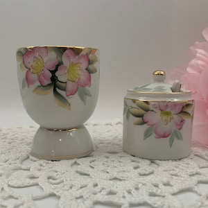 May include: A white ceramic egg cup and a small white ceramic jar with lids, both decorated with pink and yellow flowers and gold trim. The items are sitting on a white crocheted doily.