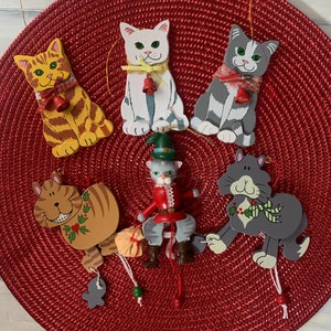 May include: Six wooden cat ornaments with bells and painted details. The cats are in various poses and colors, including gray, white, and orange. The ornaments are hanging from a red crocheted placemat.