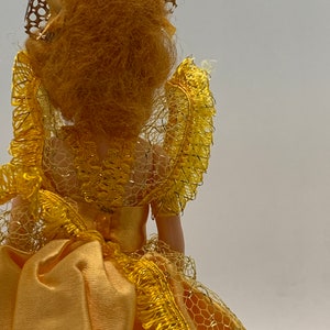 Op de afbeelding: Een vintage pop die een gele jurk draagt met een kanten overlay en een gouden kroon. De pop heeft lang, krullend, blond haar.