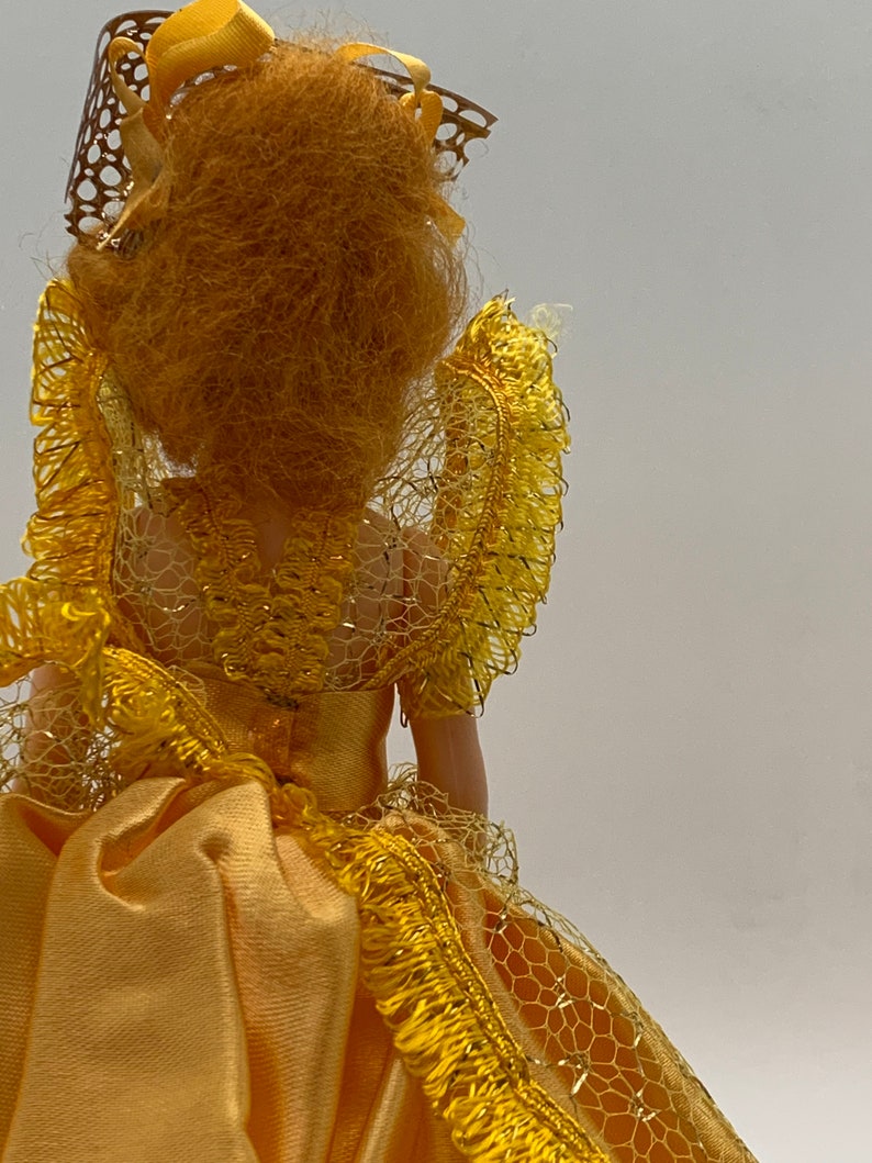 Op de afbeelding: Een vintage pop die een gele jurk draagt met een kanten overlay en een gouden kroon. De pop heeft lang, krullend, blond haar.