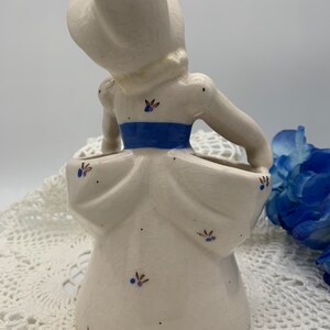 May include: A white ceramic figurine of a woman in a blue and white dress. The figurine is a planter with a small opening in the back for flowers. The figurine has a crackled glaze and is decorated with small blue and brown flowers.