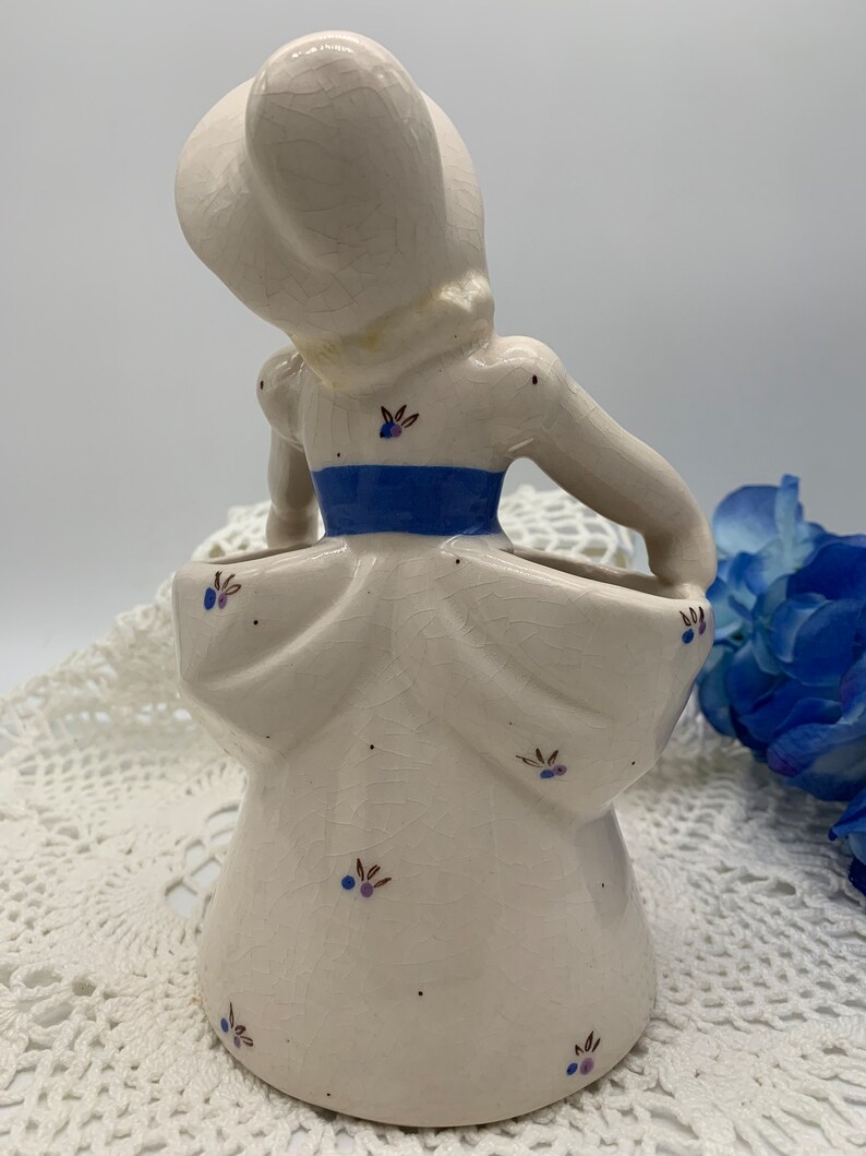 May include: A white ceramic figurine of a woman in a blue and white dress. The figurine is a planter with a small opening in the back for flowers. The figurine has a crackled glaze and is decorated with small blue and brown flowers.