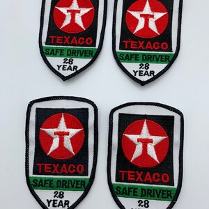 May include: Four white fabric patches with black borders. Each patch features a red star with a white outline and the text "TEXACO" in white. Below the star, each patch has a green stripe with the text "SAFE DRIVER" in white. Below the green stripe, each patch has the text "28 YEAR" in white.