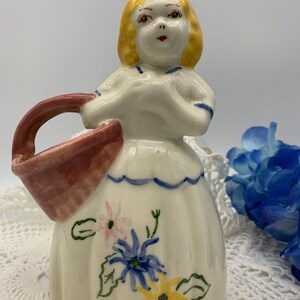 May include: A ceramic figurine of a girl in a white dress with blue trim and floral accents. She is holding a brown basket with a handle. The figurine is painted with yellow, blue, pink, and green details.