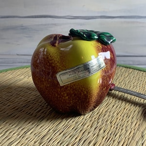 May include: A ceramic apple-shaped jar with a yellow and red glaze. The jar has a green leaf on top and a label that reads "100% Pure U.S. Grade A Honey". A small metal spoon rests on the side. The jar is on a woven mat.