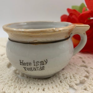 May include: White ceramic cup with a brown rim and a gold-colored glaze. The cup has a handle and the words "Here is my regards" written on the front.
