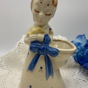 May include: A ceramic figurine of a girl with closed eyes holding a yellow bird. She is wearing a white dress with blue trim and a blue bow. The figurine has a small bowl attached to the side.