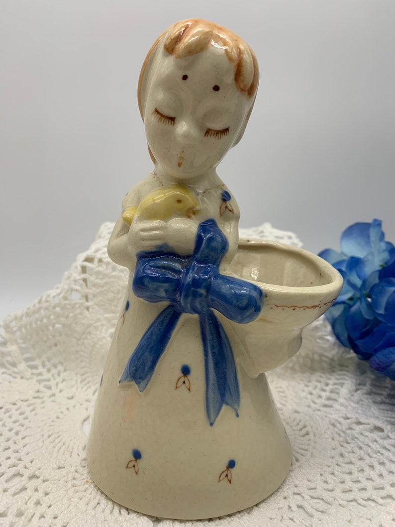 May include: A ceramic figurine of a girl with closed eyes holding a yellow bird. She is wearing a white dress with blue trim and a blue bow. The figurine has a small bowl attached to the side.