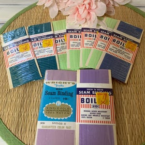 May include: A collection of vintage seam binding packages in various colors, including teal, green, and lavender. The packages feature text and are arranged on a woven mat with pink flowers in the background. Each package contains 3 yards of rayon.