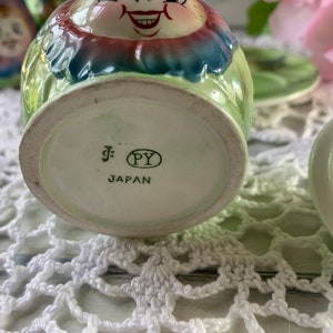 May include: A green and white ceramic sugar bowl with a smiling face and floral design. The bottom of the bowl has a label that reads "J. PY JAPAN".
