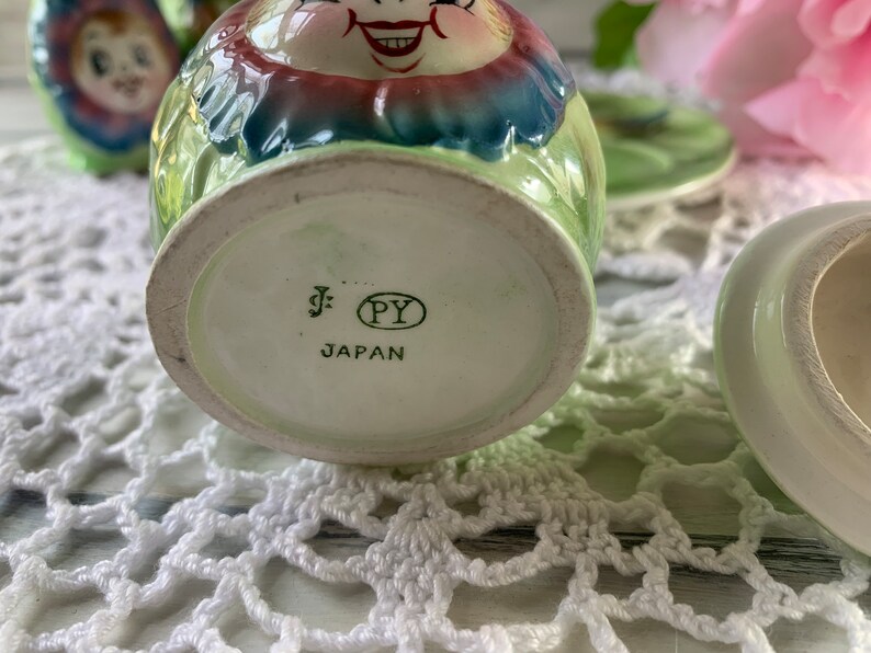 May include: A green and white ceramic sugar bowl with a smiling face and floral design. The bottom of the bowl has a label that reads "J. PY JAPAN".