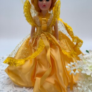 Op de afbeelding: Een kleine pop die een gele satijnen jurk draagt met een kant-overlay en een gouden kroon. De pop heeft blond haar en blauwe ogen.