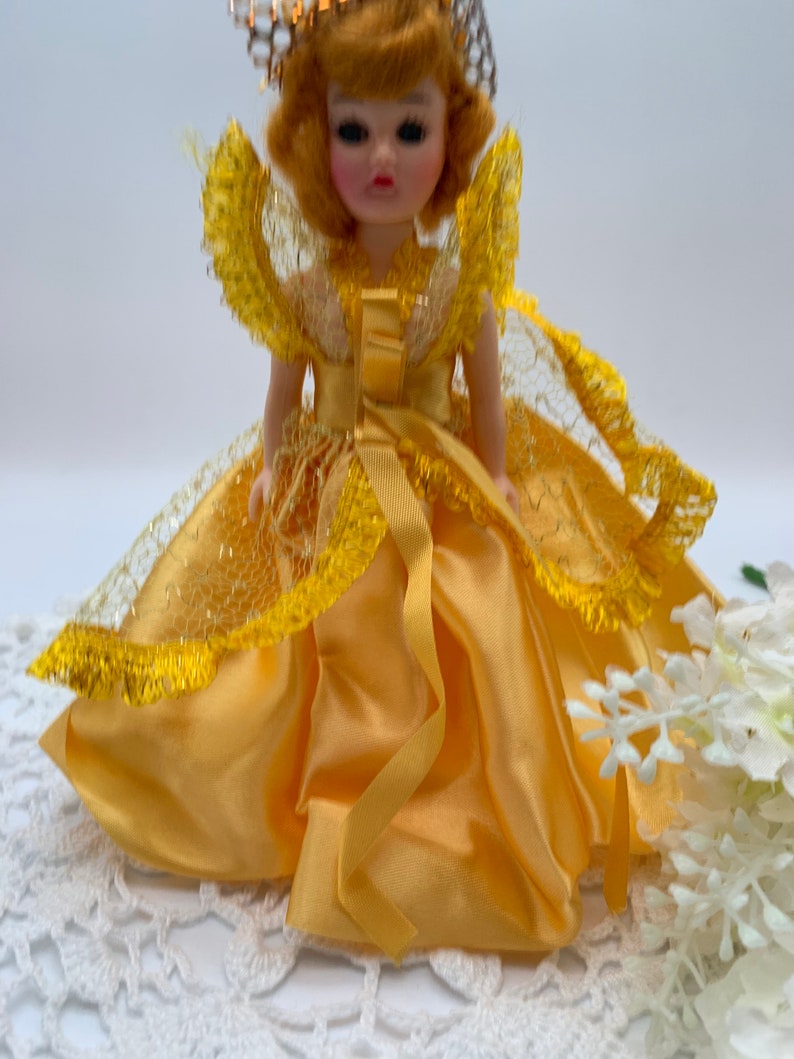 Op de afbeelding: Een kleine pop die een gele satijnen jurk draagt met een kant-overlay en een gouden kroon. De pop heeft blond haar en blauwe ogen.