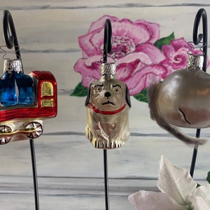 May include: Three vintage glass Christmas ornaments on black metal stands. The ornaments are a red and blue train, a silver dog, and a silver mouse with a gray fur tail.