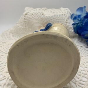 May include: A white ceramic vase with a blue ribbon around the neck. The vase has a round base and is sitting on a white crocheted surface.