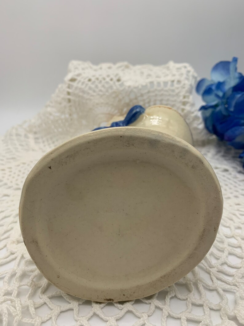 May include: A white ceramic vase with a blue ribbon around the neck. The vase has a round base and is sitting on a white crocheted surface.