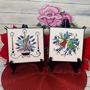 May include: Two vintage Christmas-themed ceramic tiles. One tile features a candle holder with three red candles and a green and white floral arrangement. The other tile features two blue and green bells with red berries and green holly leaves.