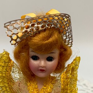 Op de afbeelding: Een vintage pop met blond haar, gekleed in een gele jurk met een kant-overlay en een zilveren en gele netmuts.