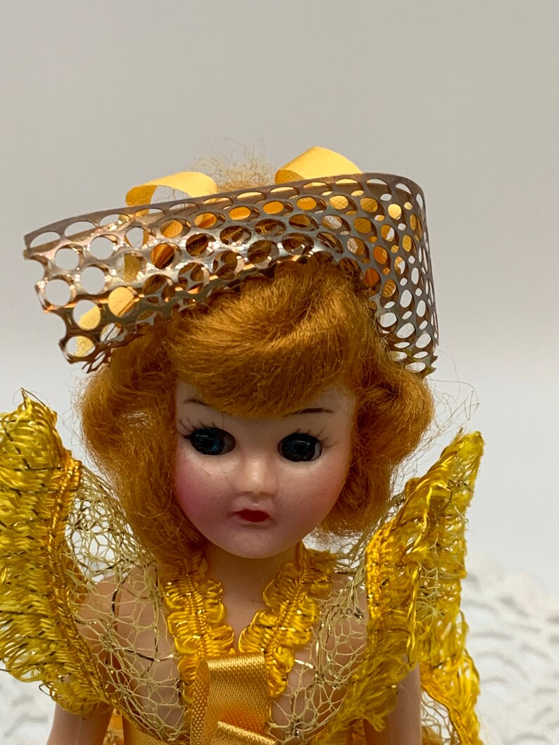 Op de afbeelding: Een vintage pop met blond haar, gekleed in een gele jurk met een kant-overlay en een zilveren en gele netmuts.