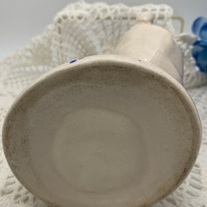 May include: White ceramic pitcher with blue and red accents. The pitcher has a rounded body and a spout. The bottom of the pitcher is visible.