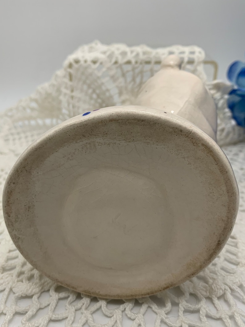 May include: White ceramic pitcher with blue and red accents. The pitcher has a rounded body and a spout. The bottom of the pitcher is visible.
