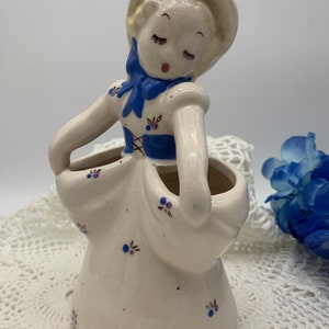May include: A white ceramic figurine of a girl with blue eyes and a blue bow. She is wearing a white dress with blue and purple flowers. The figurine is holding a white pot with two openings.