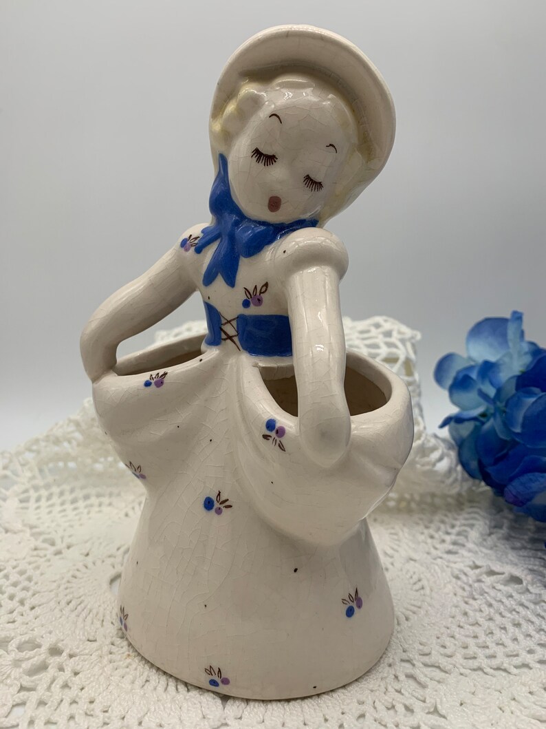 May include: A white ceramic figurine of a girl with blue eyes and a blue bow. She is wearing a white dress with blue and purple flowers. The figurine is holding a white pot with two openings.