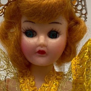 Op de afbeelding: Een vintage pop met blond haar, blauwe ogen en een gele jurk. De pop draagt een gouden kroon en een gele kanten overlay.