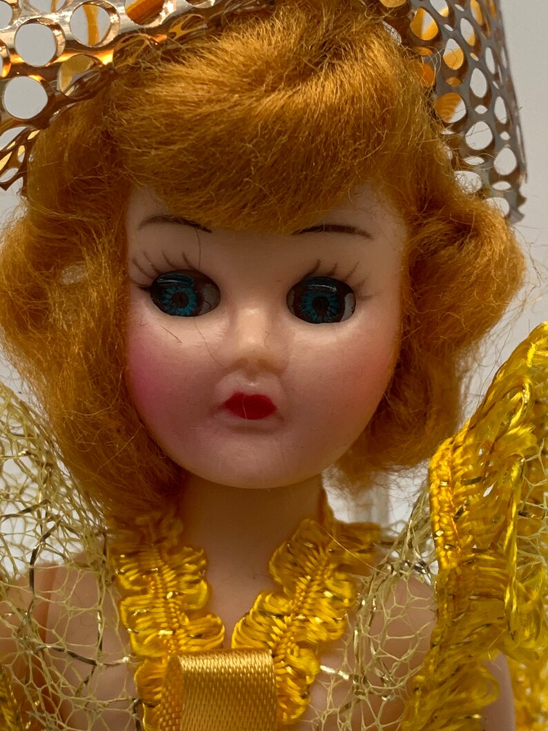 Op de afbeelding: Een vintage pop met blond haar, blauwe ogen en een gele jurk. De pop draagt een gouden kroon en een gele kanten overlay.