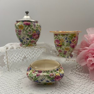 May include: Three pieces of floral patterned porcelain. A lidded jar with a silver top, a small vase, and a low bowl. The pieces are decorated with pink, yellow, and blue flowers on a white background.