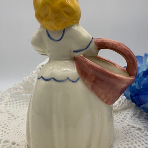 May include: A ceramic figurine of a girl in a white dress with blue trim, holding a pink and white basket. The girl has yellow hair and is wearing a blue and white collar.