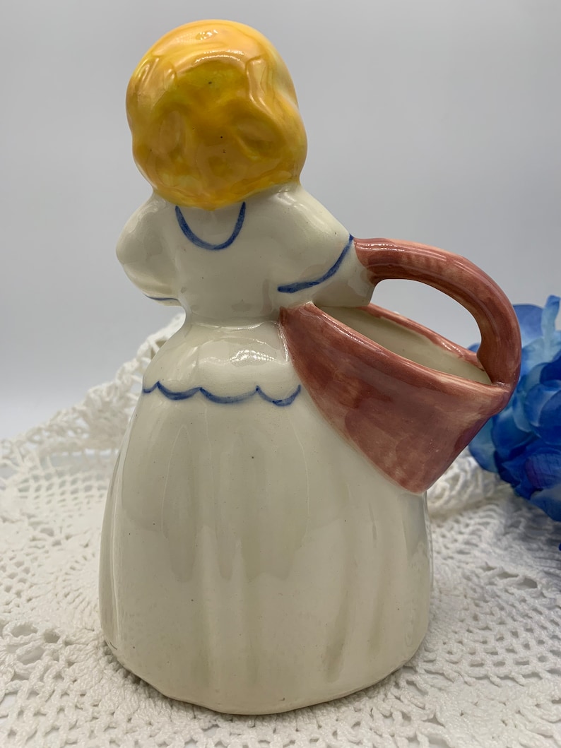 May include: A ceramic figurine of a girl in a white dress with blue trim, holding a pink and white basket. The girl has yellow hair and is wearing a blue and white collar.