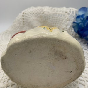 May include: The bottom of a white ceramic pitcher with a floral design. The pitcher has a rounded bottom and is decorated with yellow flowers and green leaves.