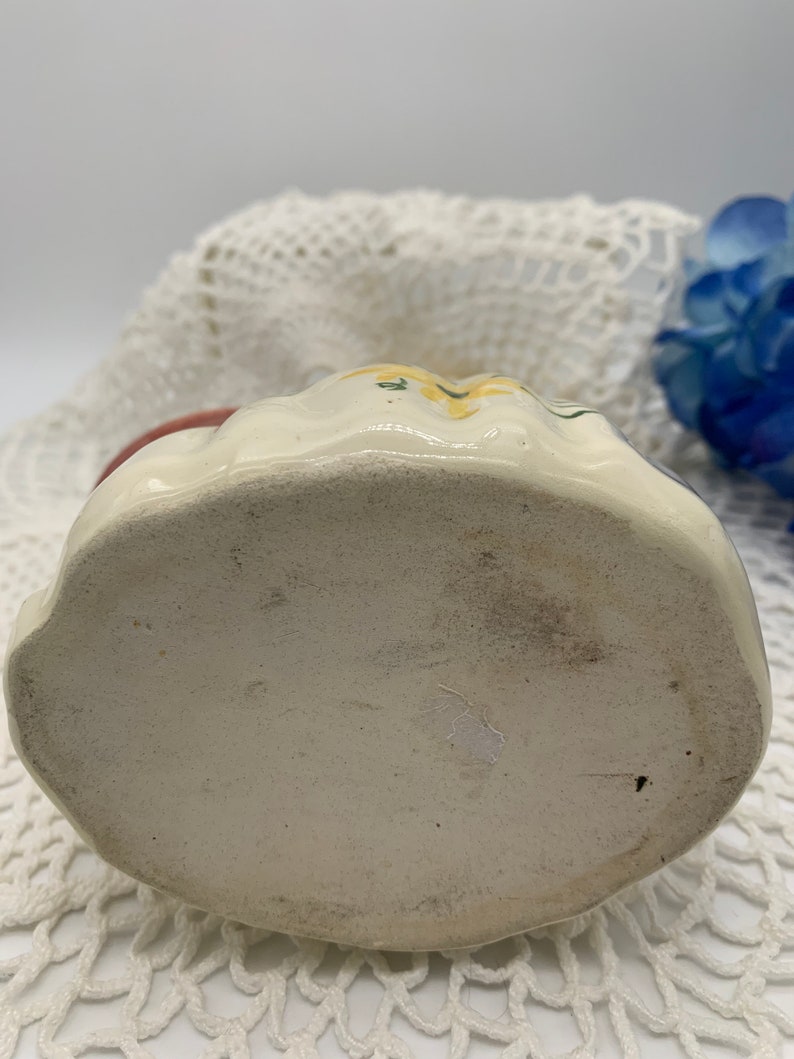 May include: The bottom of a white ceramic pitcher with a floral design. The pitcher has a rounded bottom and is decorated with yellow flowers and green leaves.