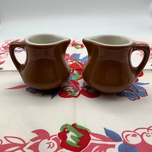 May include: Two small, brown ceramic cream pitchers with white interiors and curved handles. The pitchers are set on a floral patterned tablecloth with red, pink, green, and blue flowers.