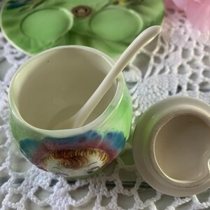 May include: A vintage ceramic sugar bowl with a green and blue floral design and a matching spoon. The bowl features a painted image of a girl with brown hair and blue eyes.