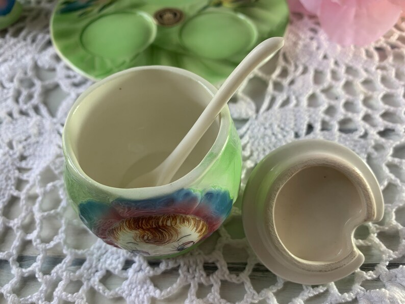 May include: A vintage ceramic sugar bowl with a green and blue floral design and a matching spoon. The bowl features a painted image of a girl with brown hair and blue eyes.