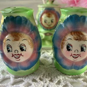 May include: Two vintage salt and pepper shakers shaped like flowers with smiling faces. The shakers are green, blue, pink, and white.