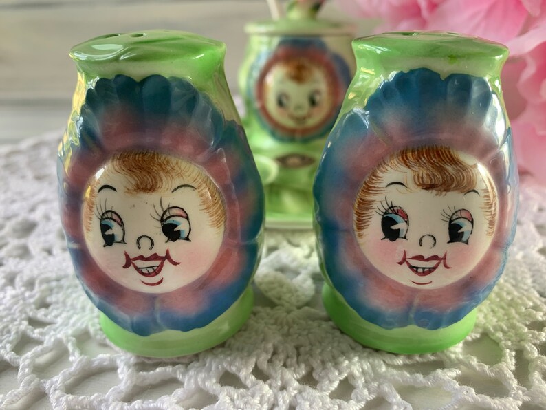May include: Two vintage salt and pepper shakers shaped like flowers with smiling faces. The shakers are green, blue, pink, and white.