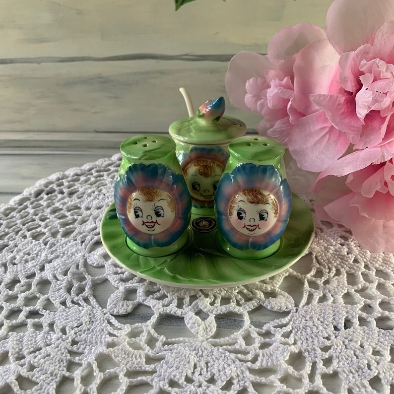 May include: A set of four green and blue ceramic salt and pepper shakers shaped like flowers with smiling faces. The shakers are on a green and white ceramic plate.