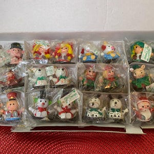 May include: A collection of small, packaged figurines in a cardboard box. The figurines depict various characters, including pigs, clowns, and snowmen, in bright colors. Some figurines have tags attached.
