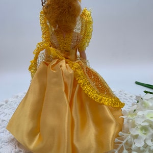 Op de afbeelding: Een gele satijnen pop met een gele kanten overlay en een bruine draadhoed. De pop draagt een gele satijnen jurk met een gele kanten overlay. De pop staat op een wit gehaakt tafellaken.