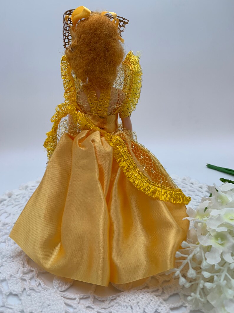 Op de afbeelding: Een gele satijnen pop met een gele kanten overlay en een bruine draadhoed. De pop draagt een gele satijnen jurk met een gele kanten overlay. De pop staat op een wit gehaakt tafellaken.