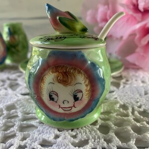 May include: A green and blue ceramic sugar bowl with a floral design and a smiling child's face. The lid has a green and pink flower design and the text "It's a Pretty Little Thing".