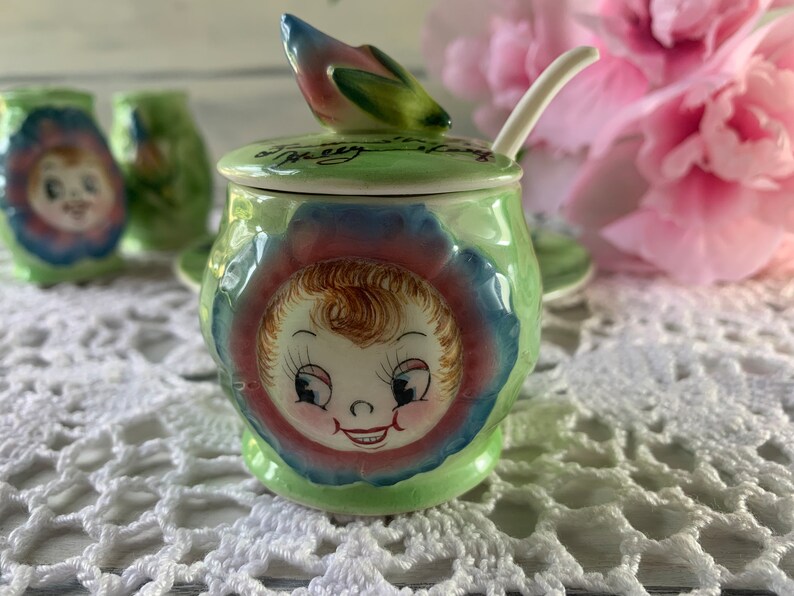 May include: A green and blue ceramic sugar bowl with a floral design and a smiling child's face. The lid has a green and pink flower design and the text "It's a Pretty Little Thing".